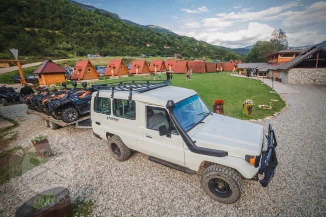 Parkirani Jeep koji koji ima prikolicu sa na kojoj su kvadovi, parkiran u kampu, u pozadini se vide bungalovi